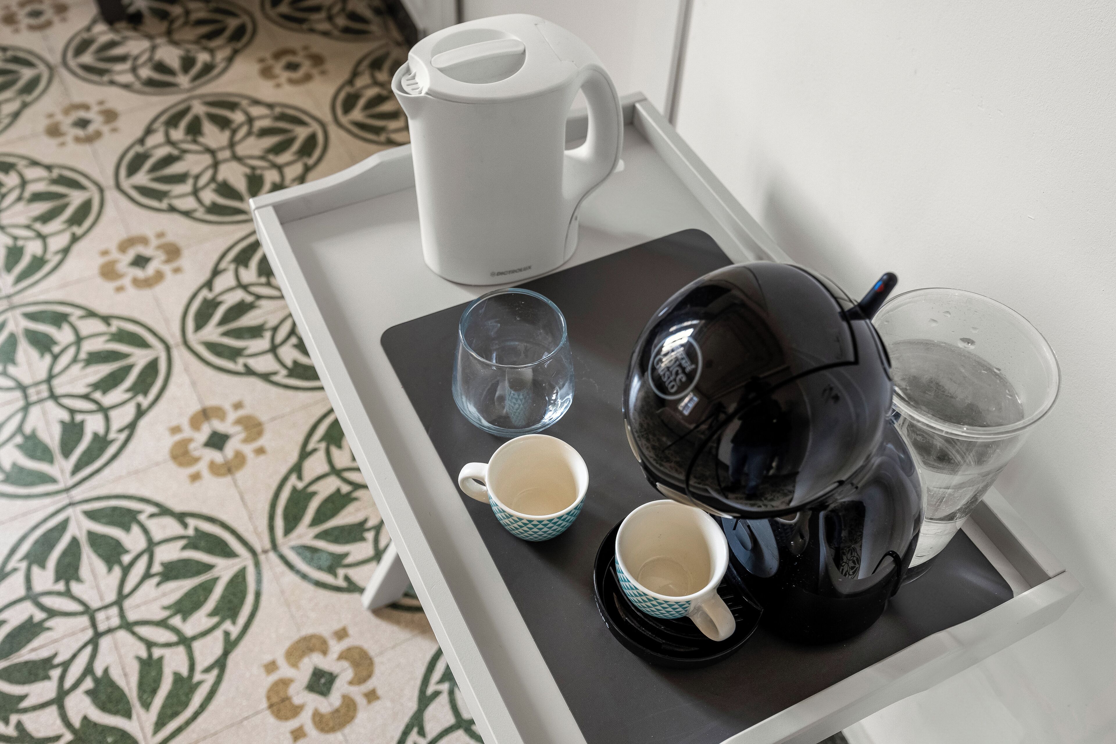 A tray with a black electric kettle, two cups, a glass, and a water jug on a patterned floor.