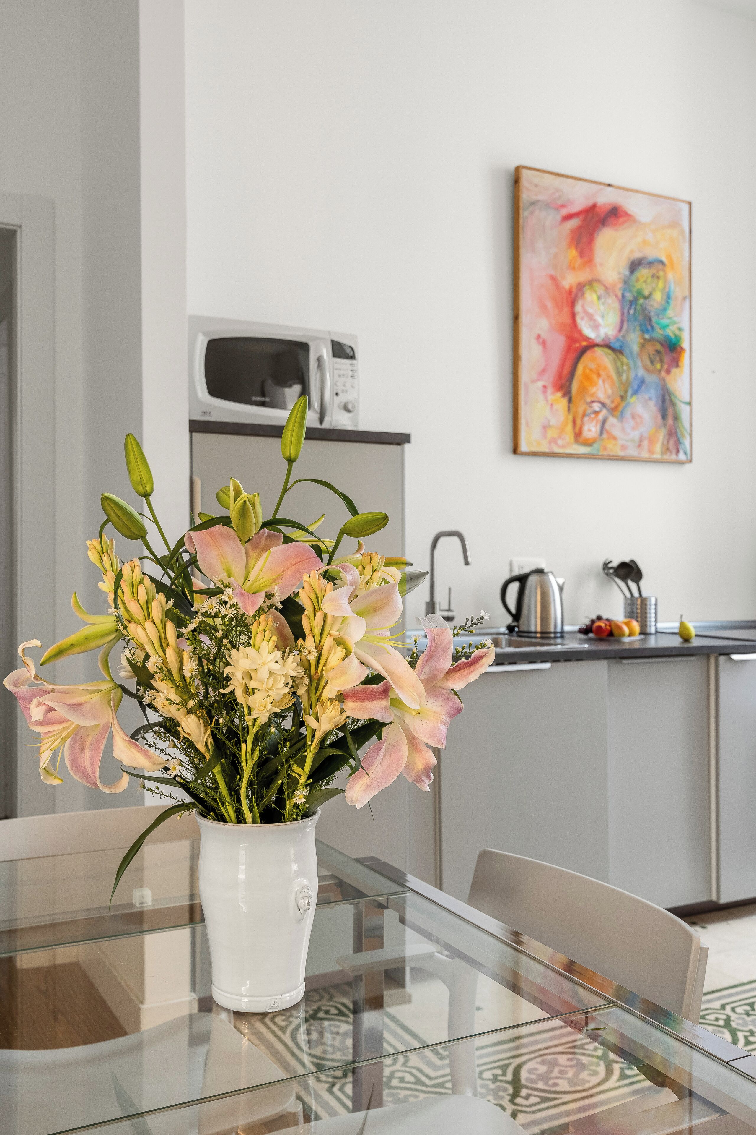A modern kitchen with a microwave, sink, and coffee maker. A white vase with flowers is on a glass table.