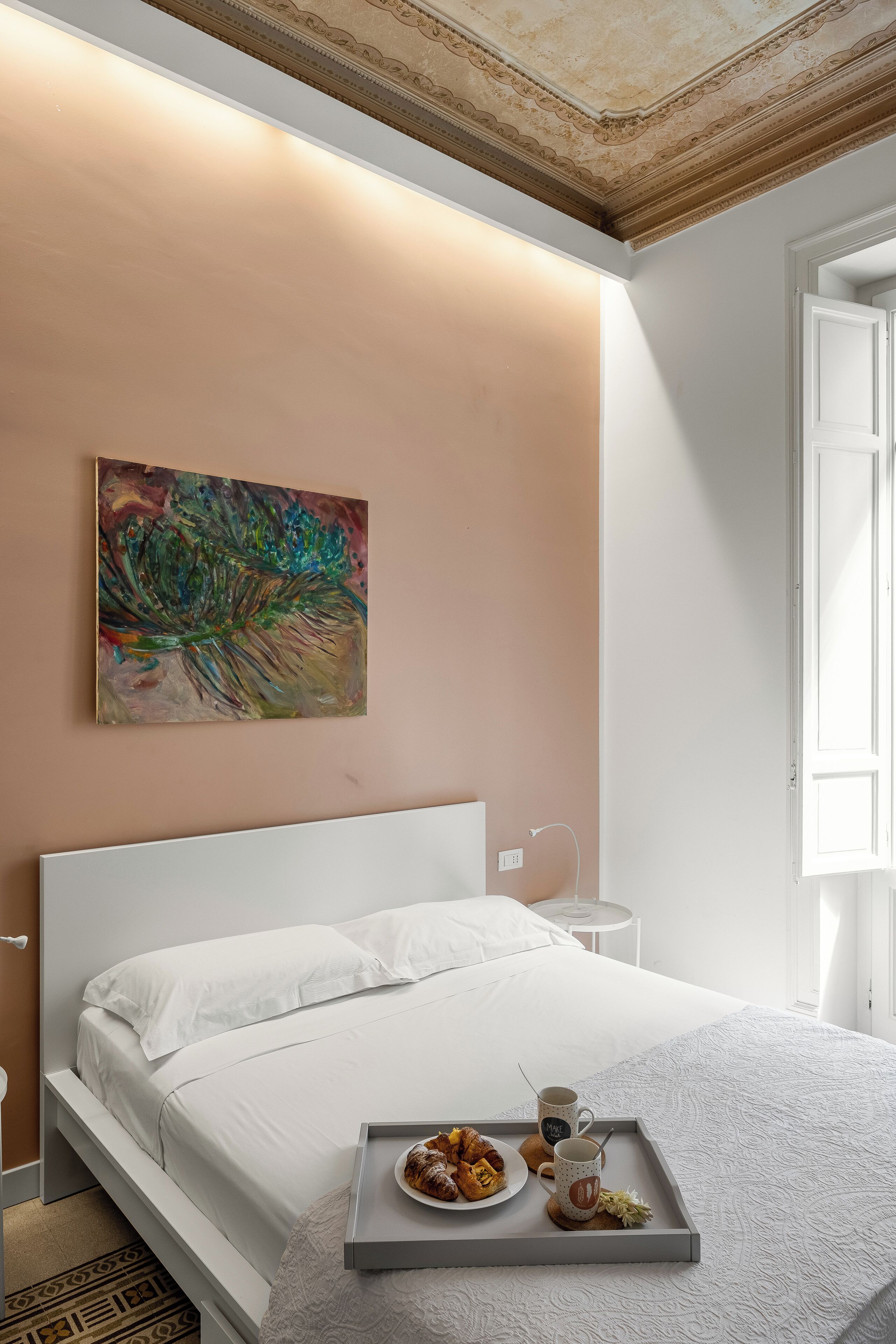 Un lit bien fait, avec un plateau de viennoiseries et des tasses de café, un tableau accroché au mur et une petite table de chevet.