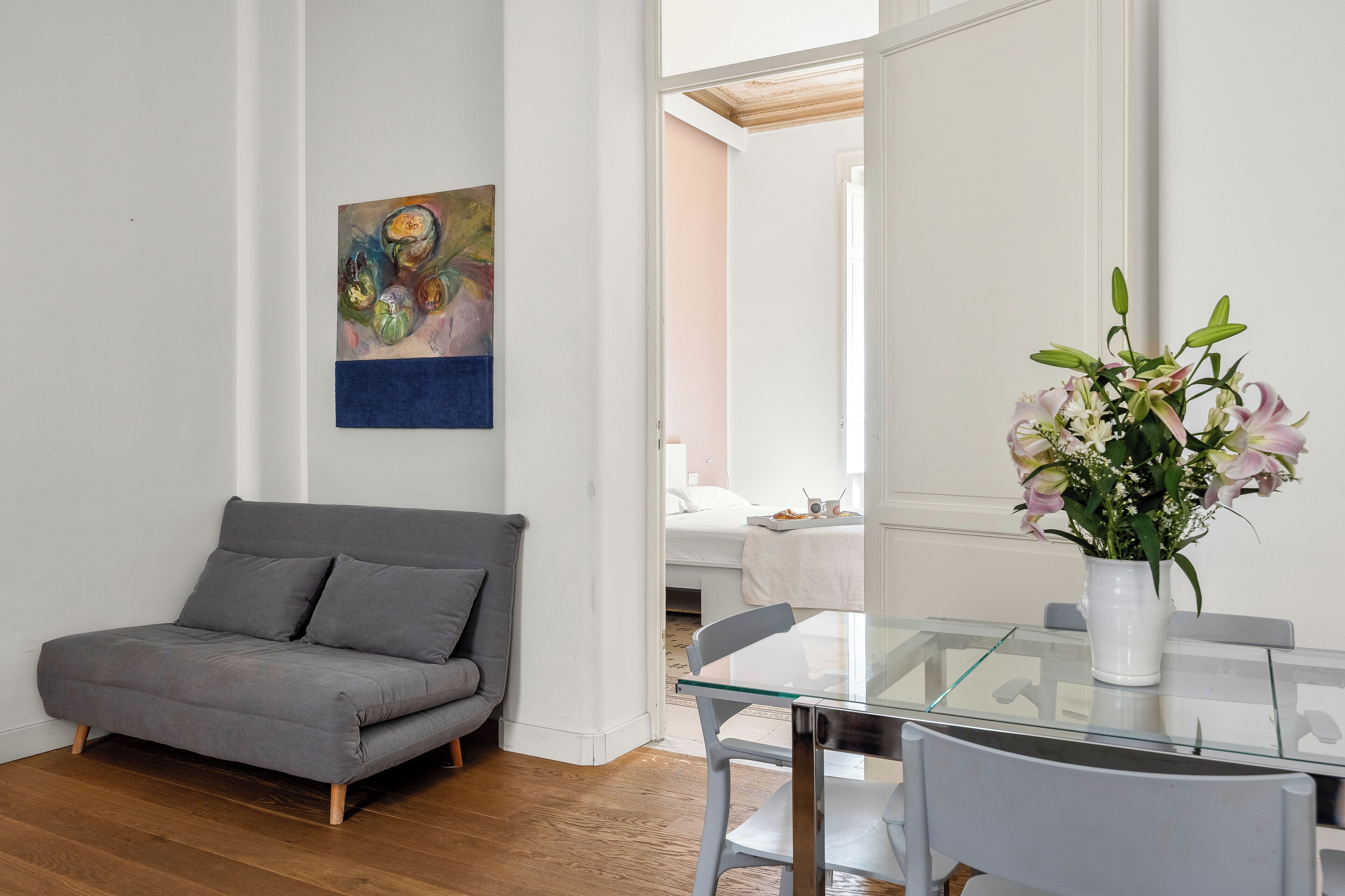 A modern living room with a grey sofa, a glass dining table, and a vase of flowers.