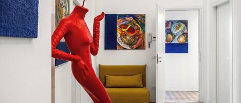 A hallway with a red mannequin, a mustard-colored sofa, and abstract paintings on the walls.