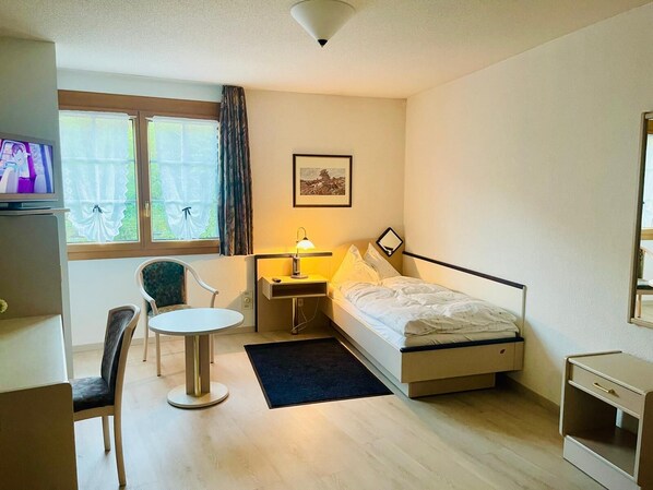 Camera d'albergo piccola con letto, comodino, lampada, sedia, scrivania e televisore.