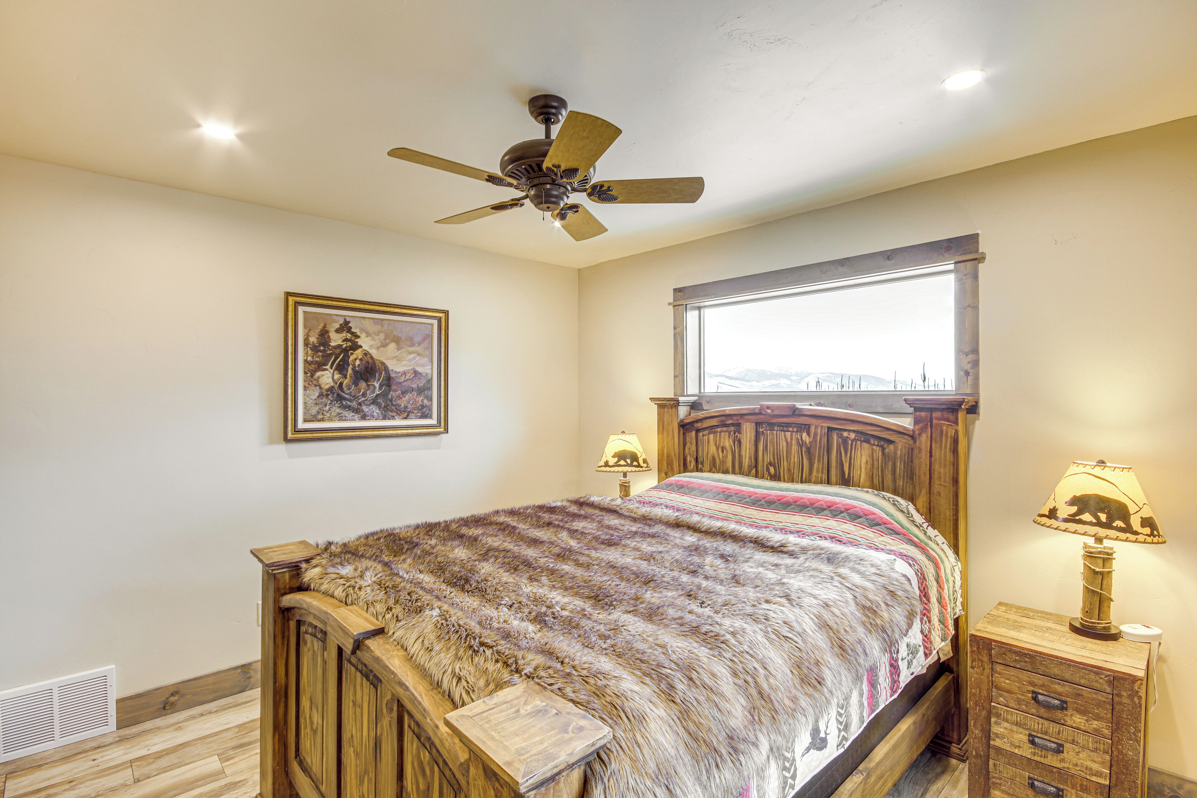 A bedroom with a wooden bed, a ceiling fan, a window with a view, a bedside lamp, and a framed picture on the wall.