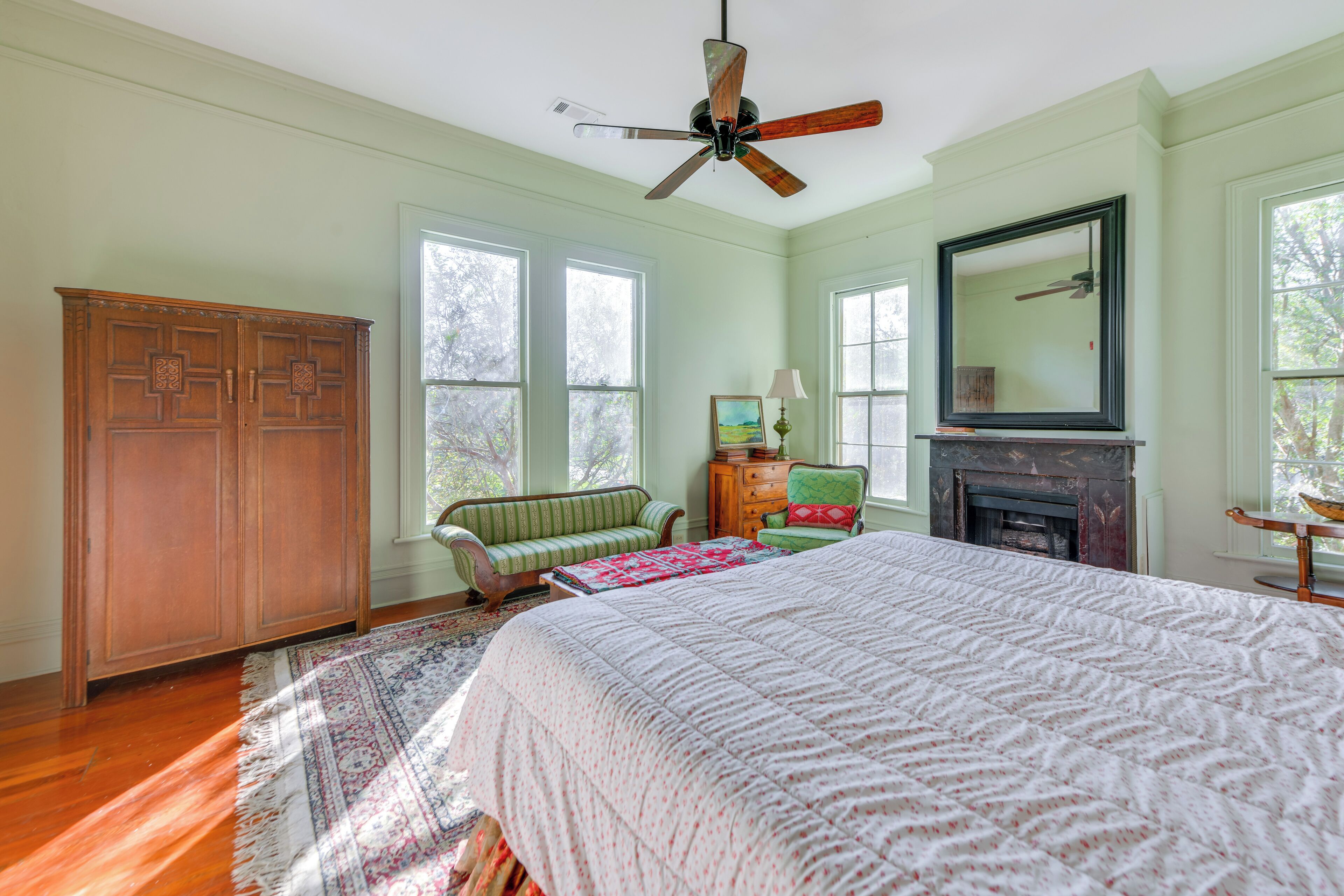 A bedroom with a large bed, a wooden wardrobe, a green armchair, a fireplace, and a ceiling fan.