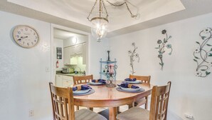 Uma área de refeições com uma mesa de madeira redonda posta para quatro pessoas, rodeada por cadeiras de madeira com assentos acolchoados. Um relógio de parede e uma peça de arte decorativa são visíveis.