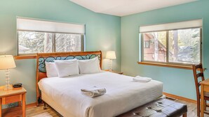 Una cama bien tendida con sábanas blancas, un cabecero de madera y un banco al pie. Hay dos mesitas de noche con lámparas y una ventana con persianas.