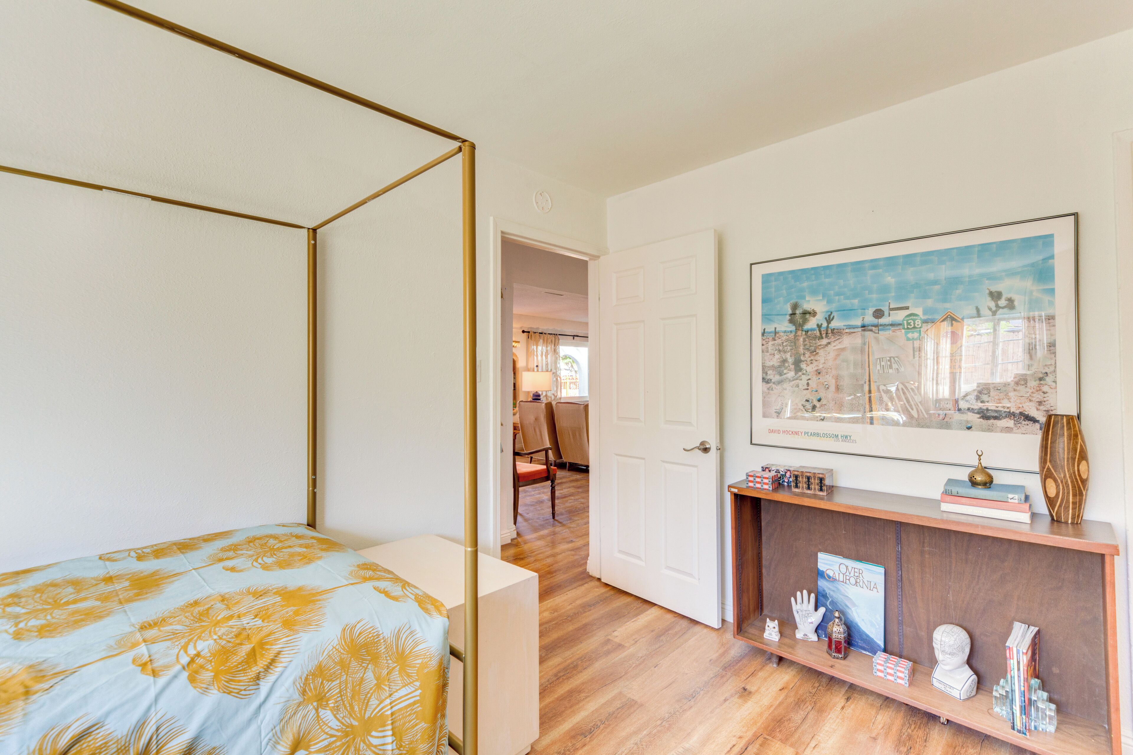 A bedroom with a bed, a door leading to another room, a shelf with books and decorative items, and a framed picture on the wall.
