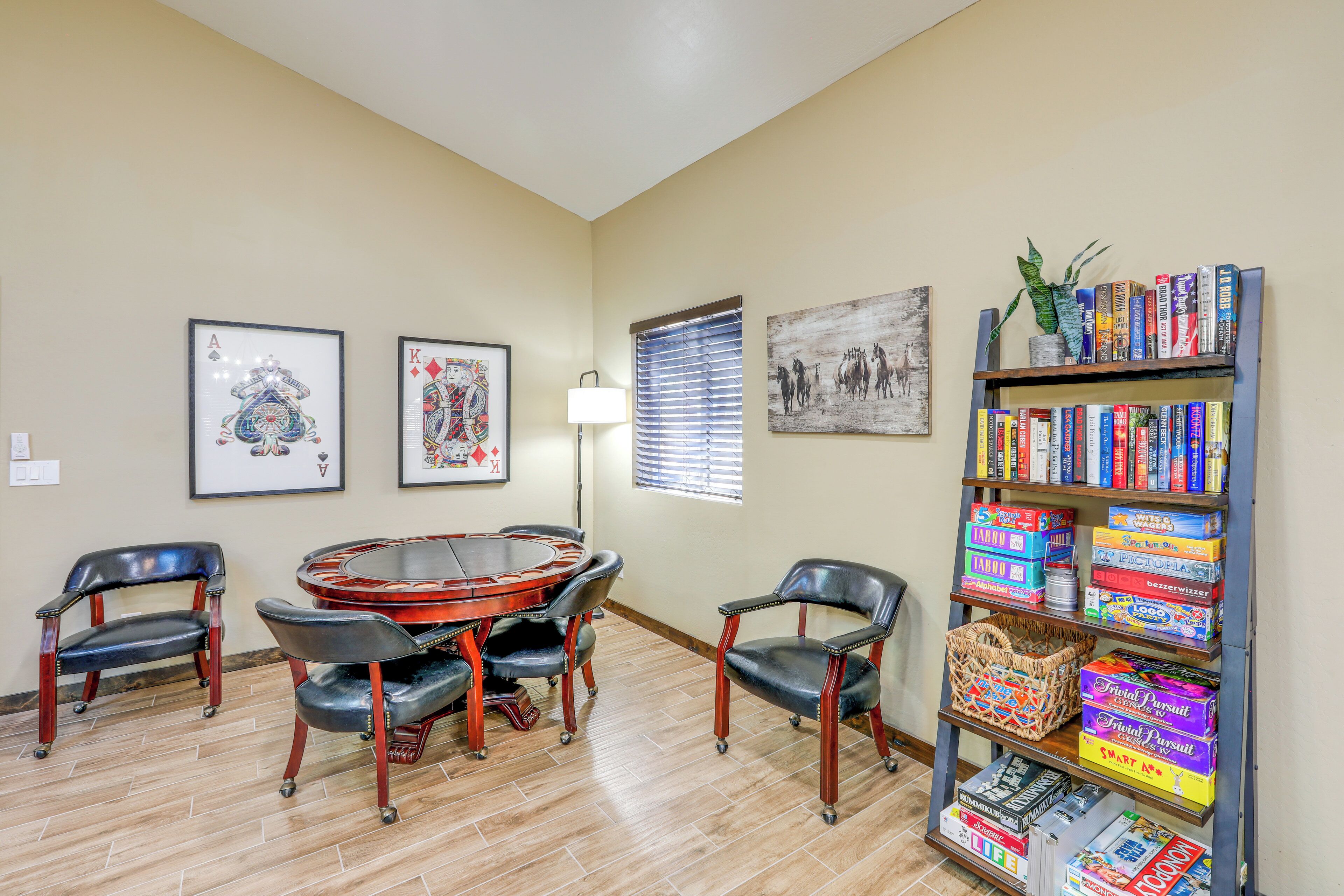A room with a round table and chairs, framed artwork on the walls, a bookshelf with books and board games, and a window with blinds.