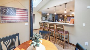 A dining area with a table, chairs, and a large American flag. There is a kitchen area with a bar and modern lighting. - Gazebo w/ View & EV Charger: Rustic Williams Cabin (Williams)