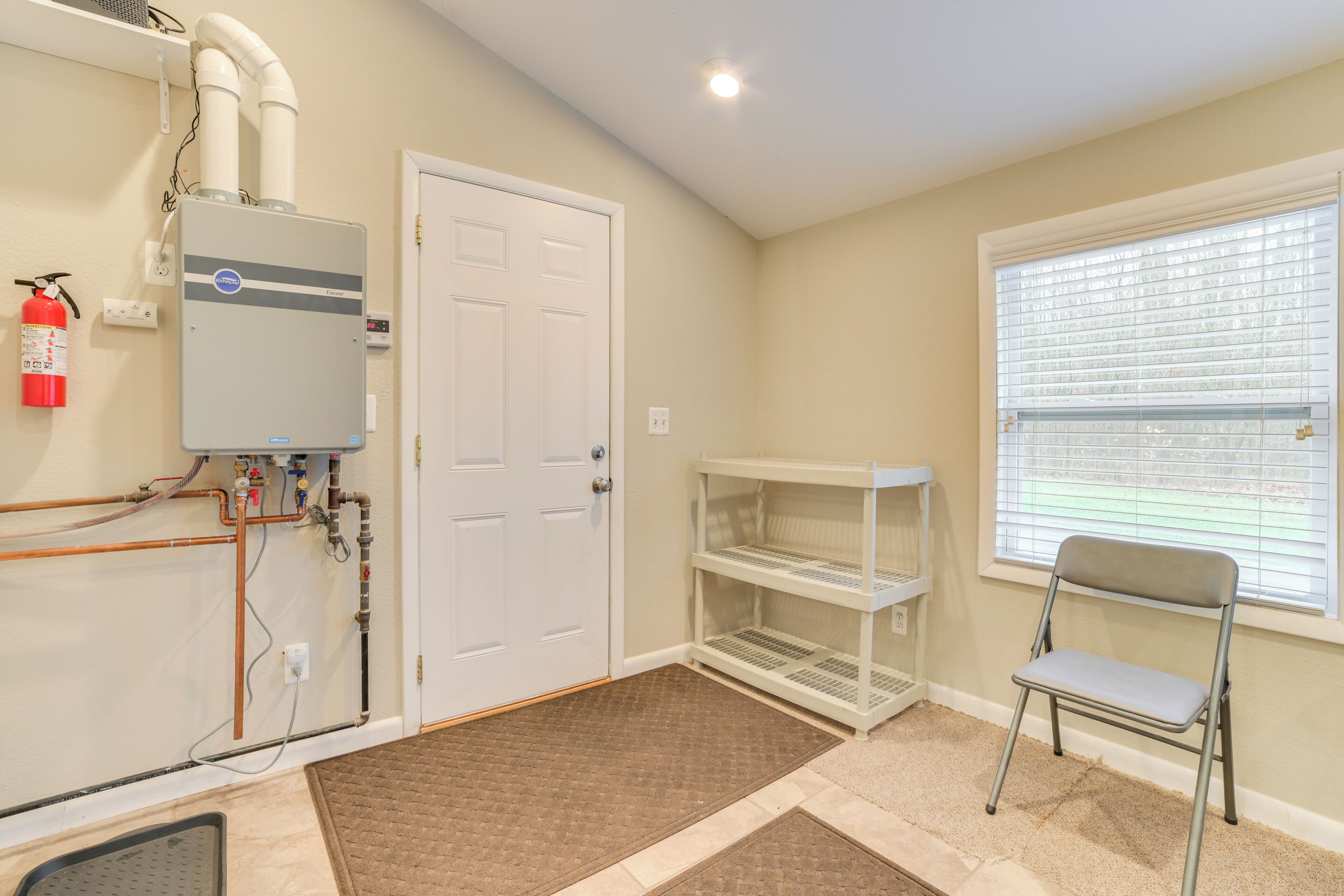 A room with a water heater, a white door, a window with blinds, and a metal shelf.