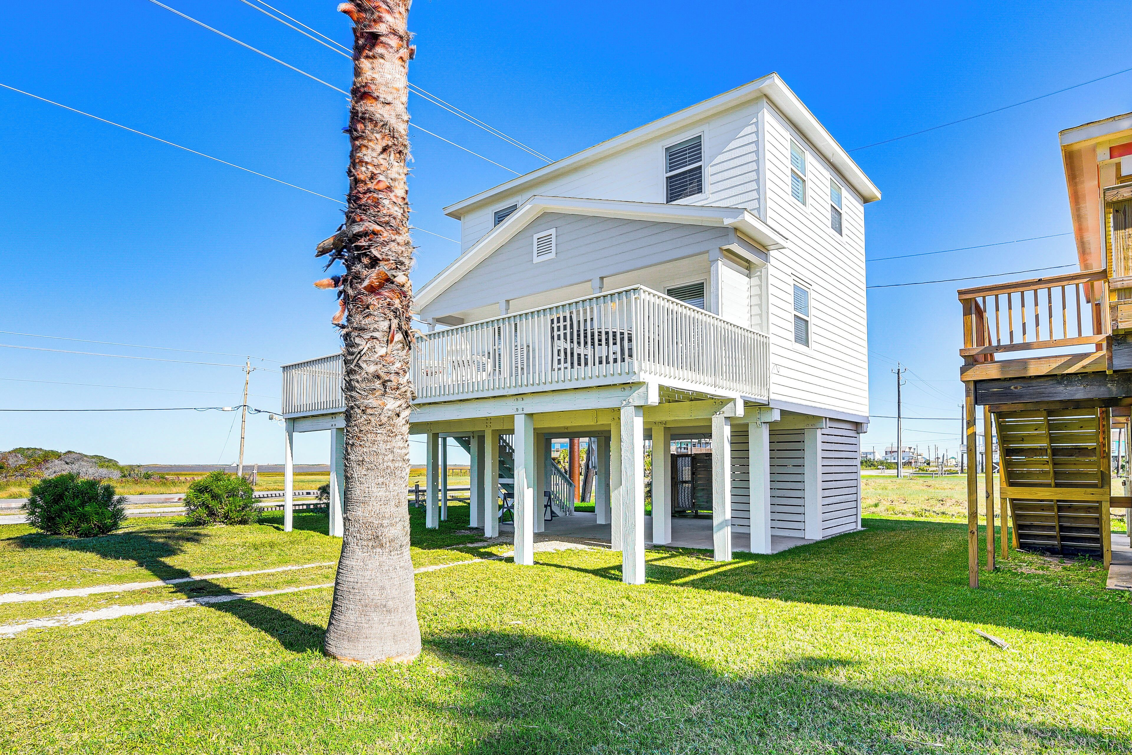 A two-story house with a porch and a balcony, surrounded by a grassy yard and a palm tree.