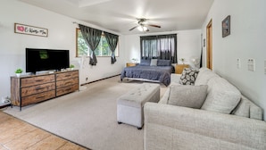 A spacious living room with a large flat-screen TV, a ceiling fan, a sofa, a bed, and a wooden cabinet. - 4 Mi to Beach: Keaau Retreat w/ Relaxing Porch! (Keaau)