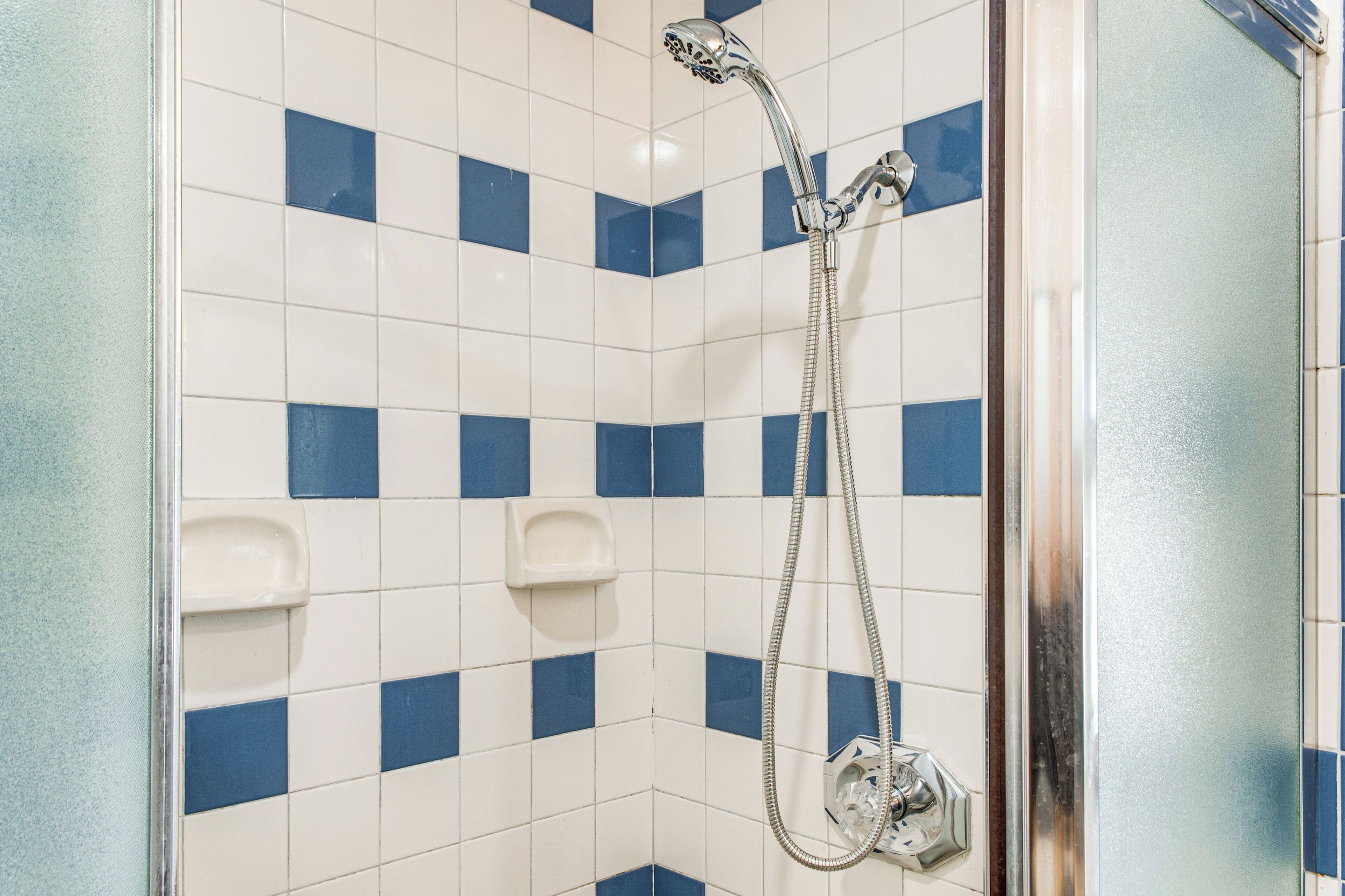 Zona de ducha con azulejos azules y blancos, un cabezal de ducha manual y un panel de control en la pared.