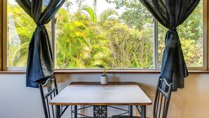 A room with a table and chairs, a window with curtains, and a view of greenery outside. - 4 Mi to Beach: Keaau Retreat w/ Relaxing Porch! (Keaau)