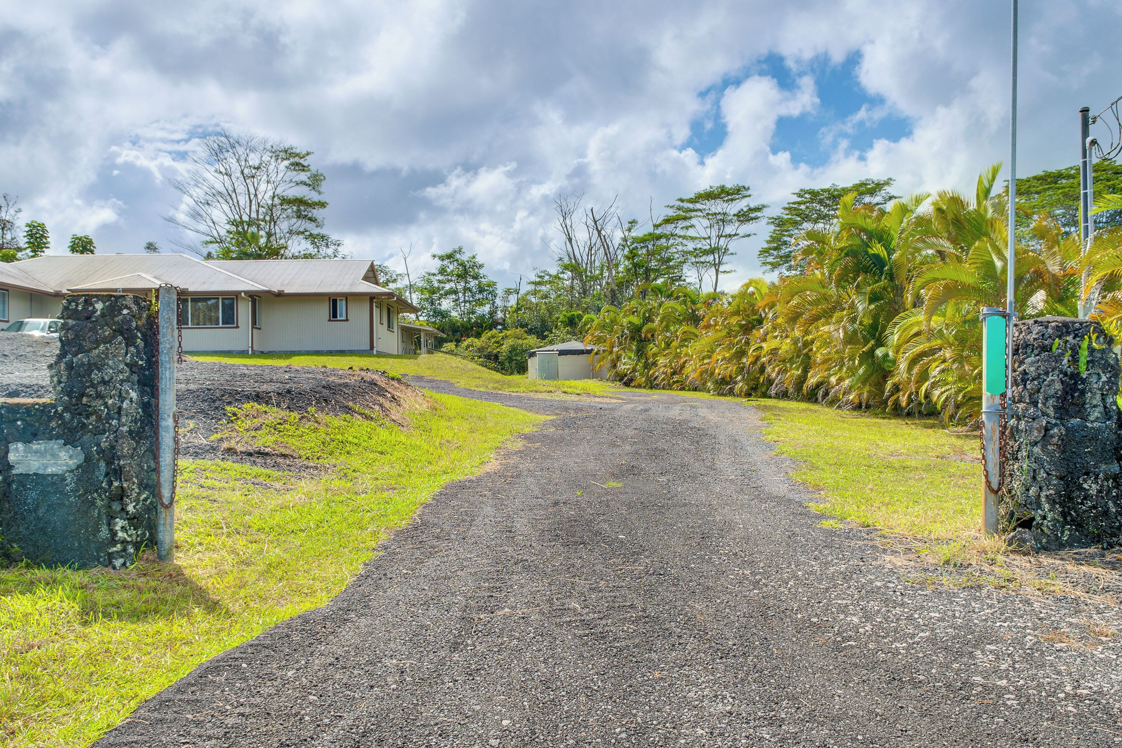 A gravel driveway leading to a house with a metal gate, surrounded by lush greenery and palm trees.