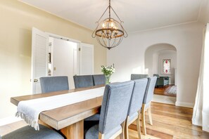 Ein Essbereich mit Holztisch, grau gepolsterten Stühlen, einer Chandelierleuchtre, und Blick auf einen Flur mit Tür und einem Buntglasfenster.