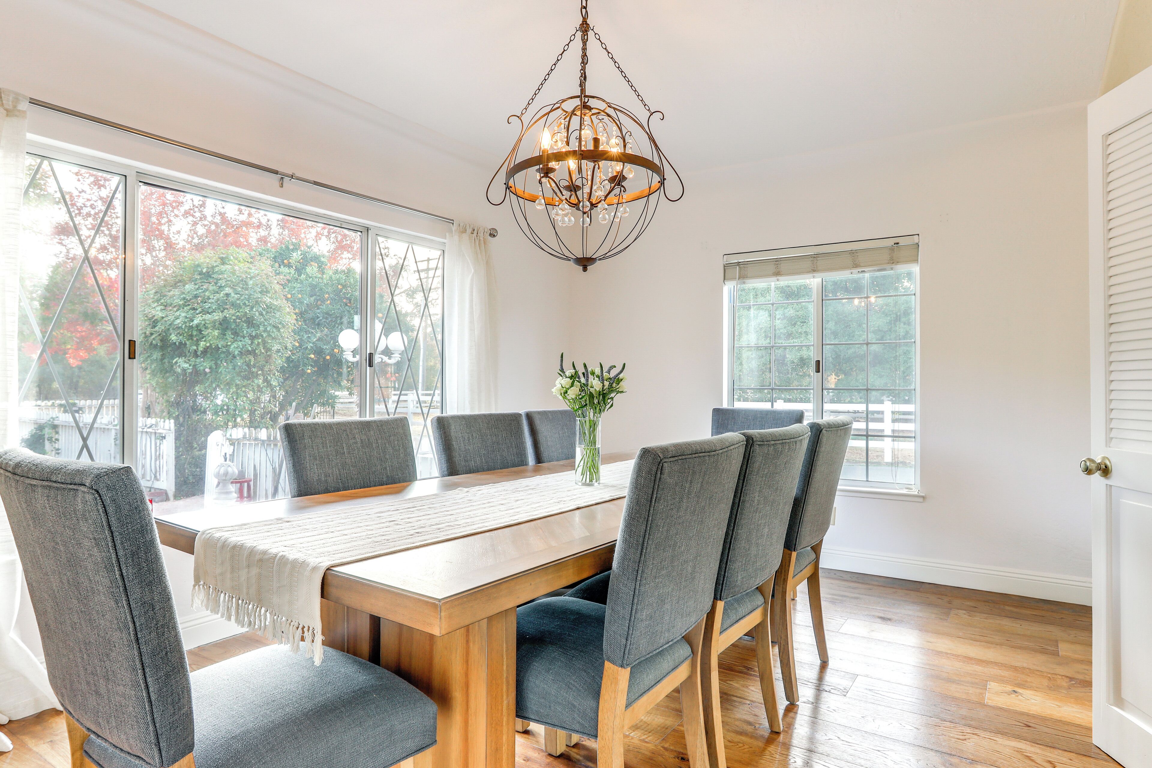 A dining room with a wooden table, grey upholstered chairs, a chandelier, and large windows with a view of trees and a fence.
