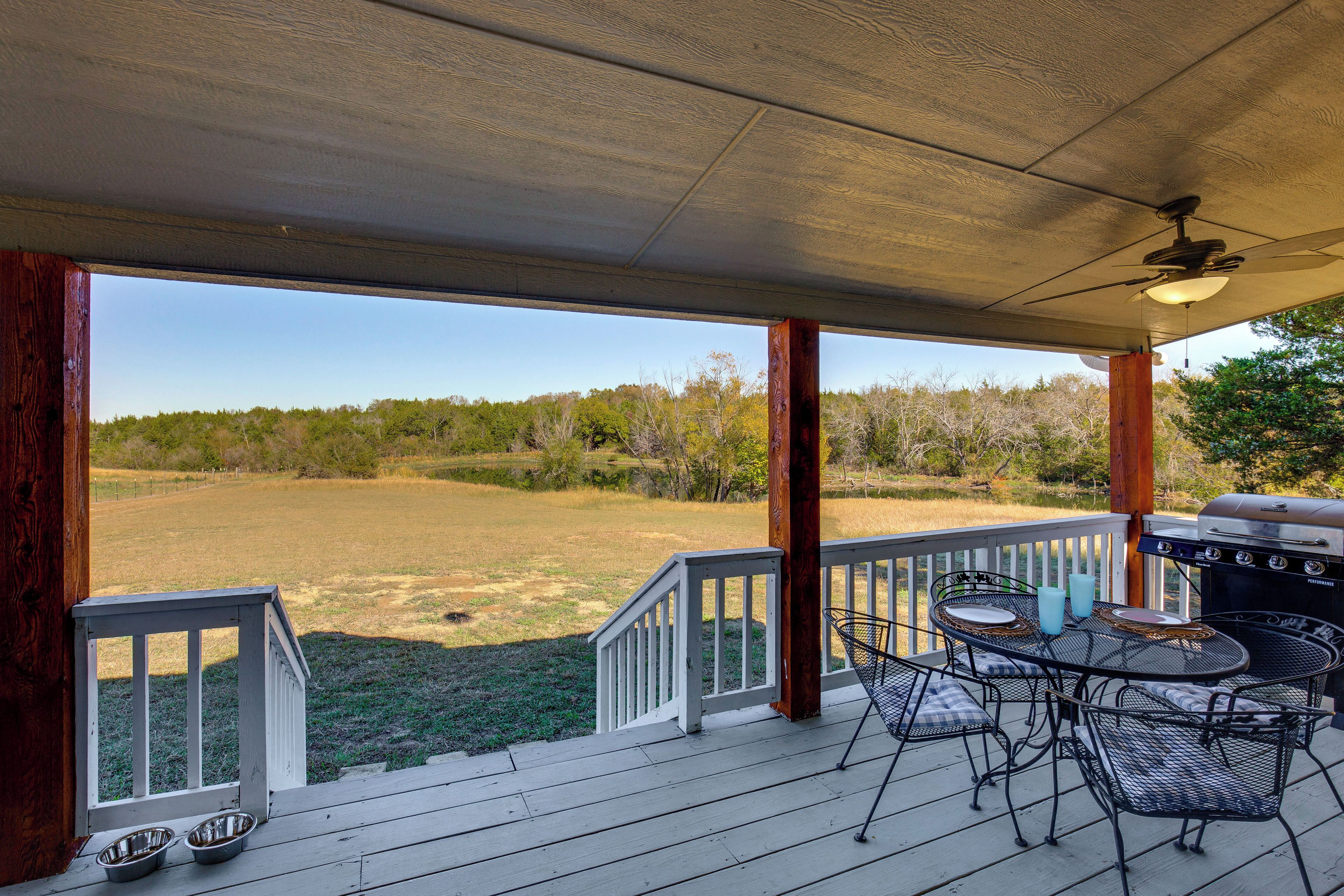 Un patio techado al aire libre con vistas a un campo de césped, una parrilla de barbacoa y una mesa con sillas.