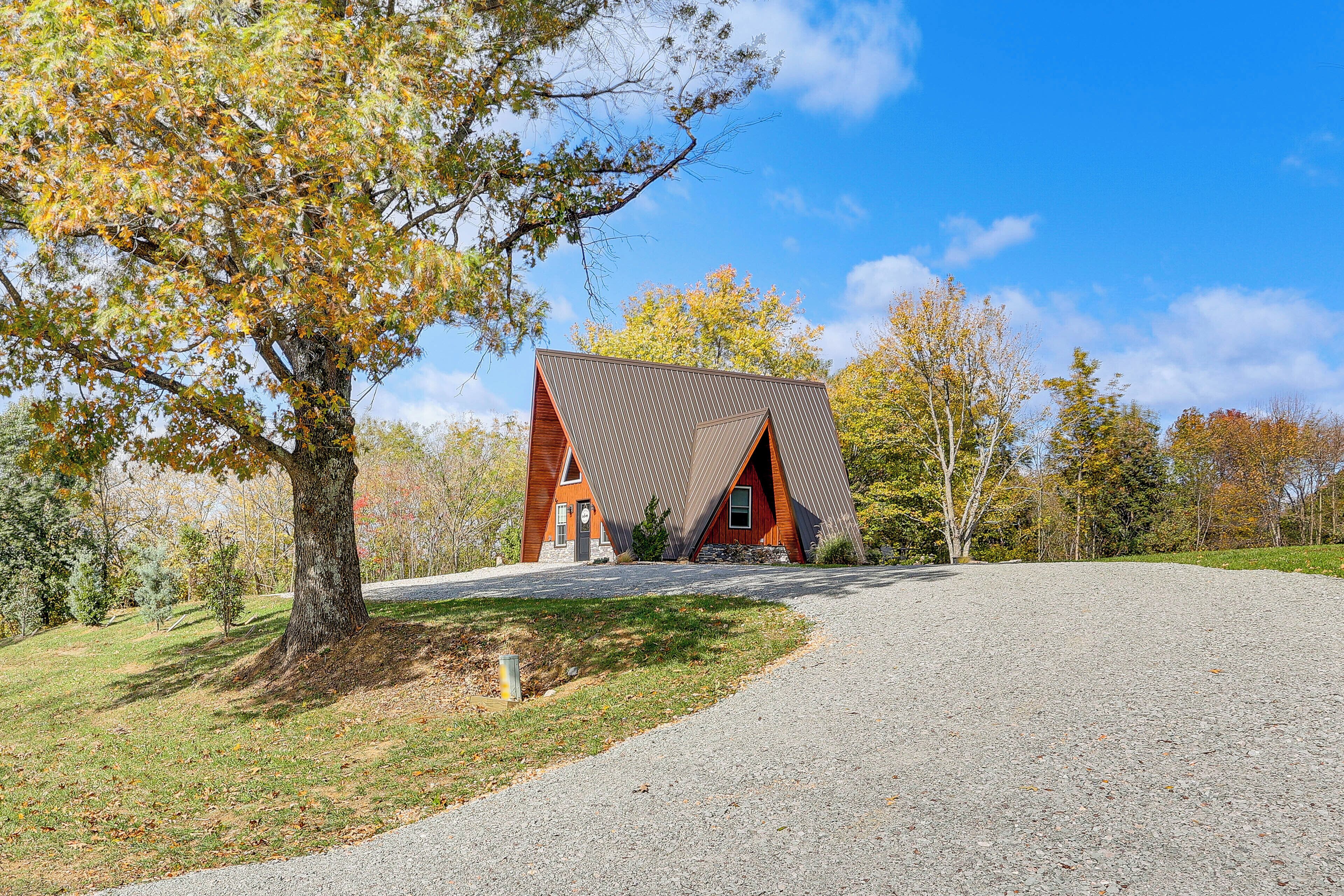 A modern house with a unique triangular roof, surrounded by trees and a gravel driveway.