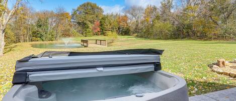 A hot tub in a backyard with a wooden deck, a pond, and trees in the background.