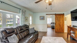 A living room with a leather sofa, wooden floor, ceiling fan, and a view through large windows. - Walk to Sandusky Bay: Home w/ Hot Tub & Fire Pit! (Sandusky)