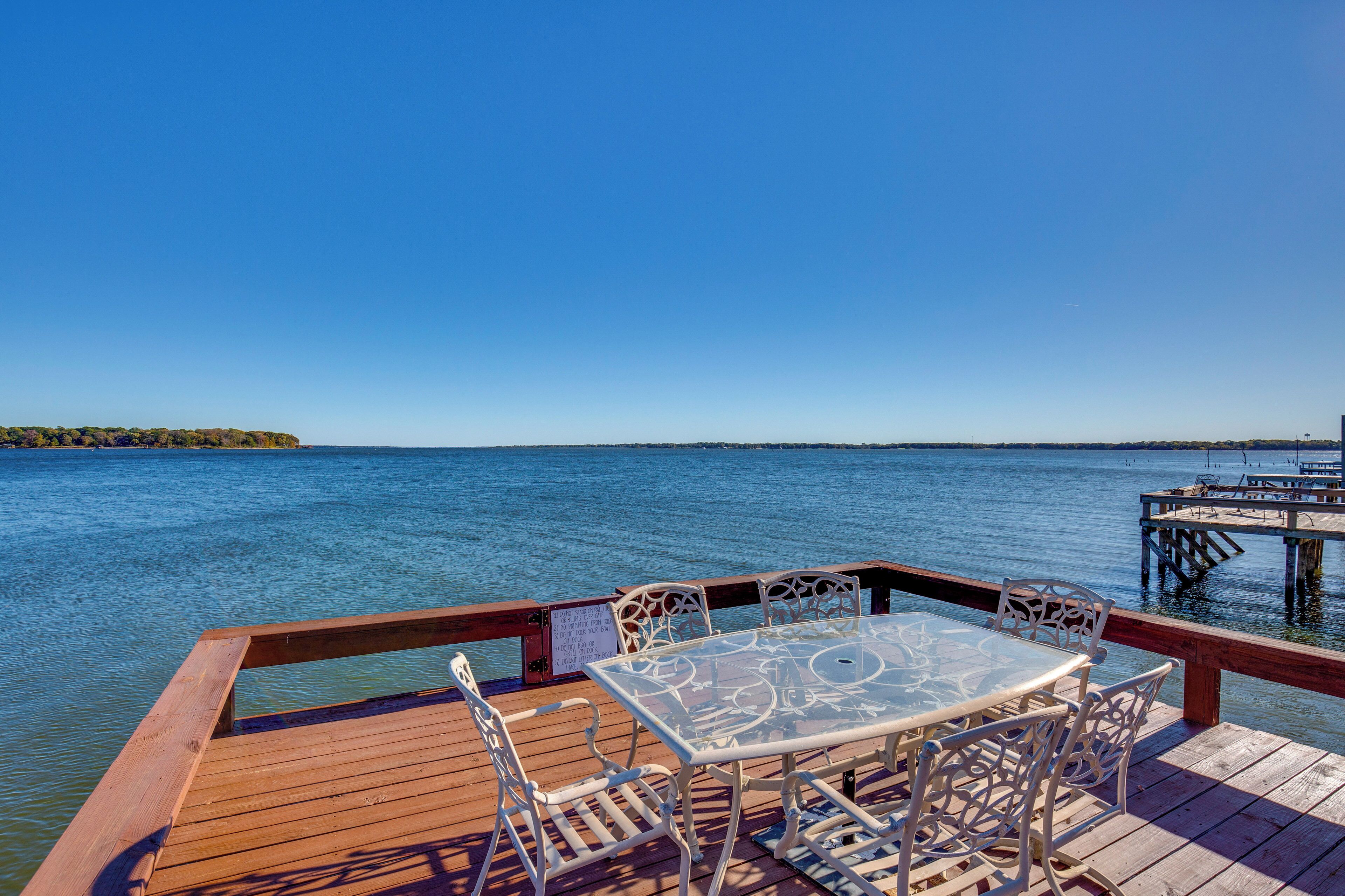 A wooden deck with a glass table and white chairs overlooking a calm body of water.