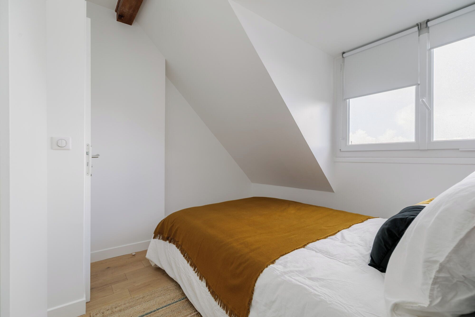 Un dormitorio con techo inclinado, una ventana con persianas, una cama con ropa blanca y un plaid marrón, y suelo de madera.