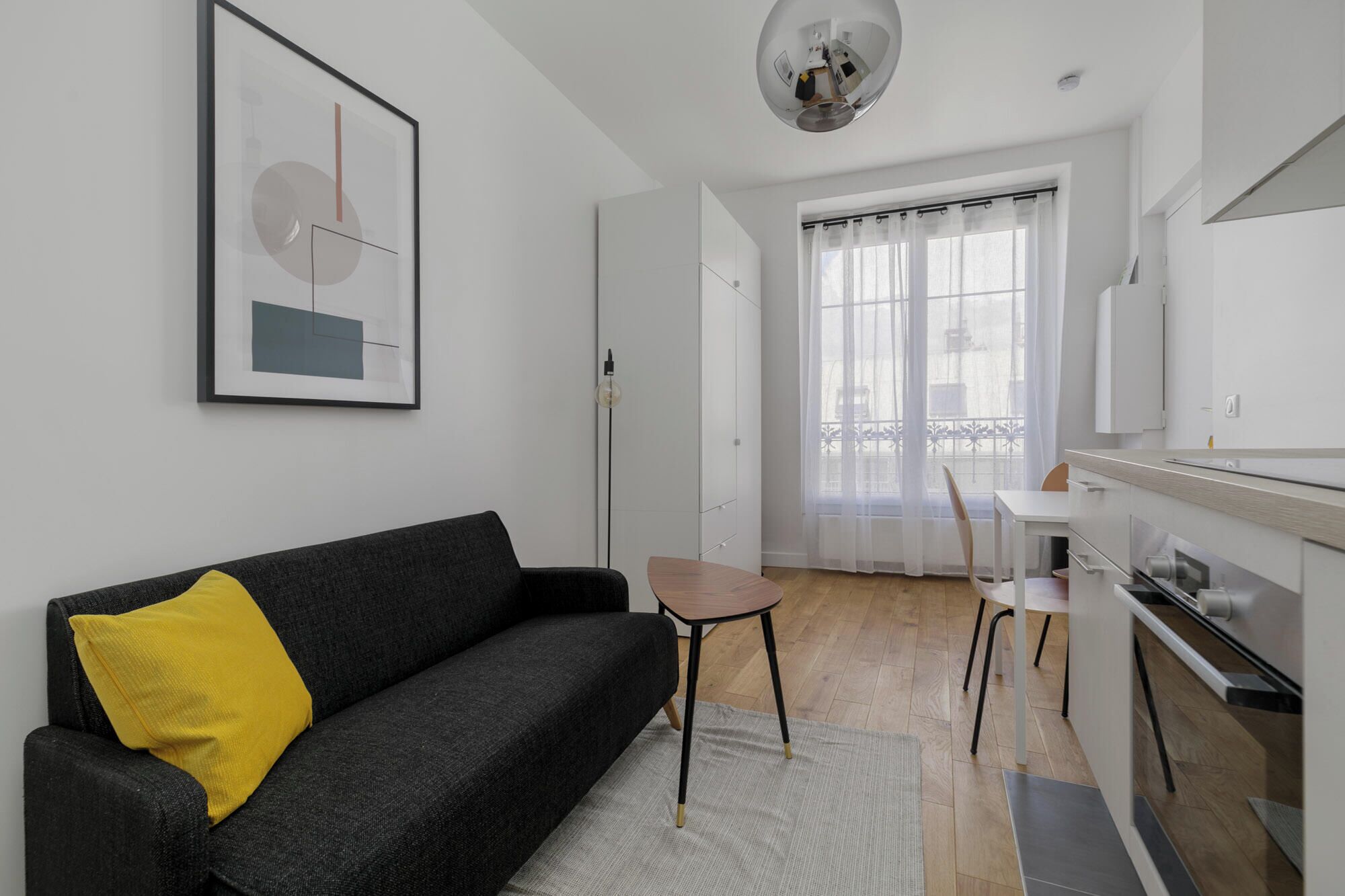 A modern living room with a black sofa, a yellow pillow, a dining area with a white table and chairs, and a kitchen with white cabinets and a stove.