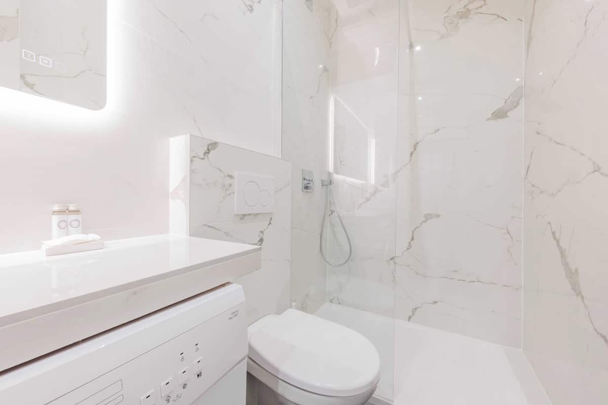 Un baño moderno con pared de mármol blanco, inodoro blanco y zona de ducha con mampara de cristal.