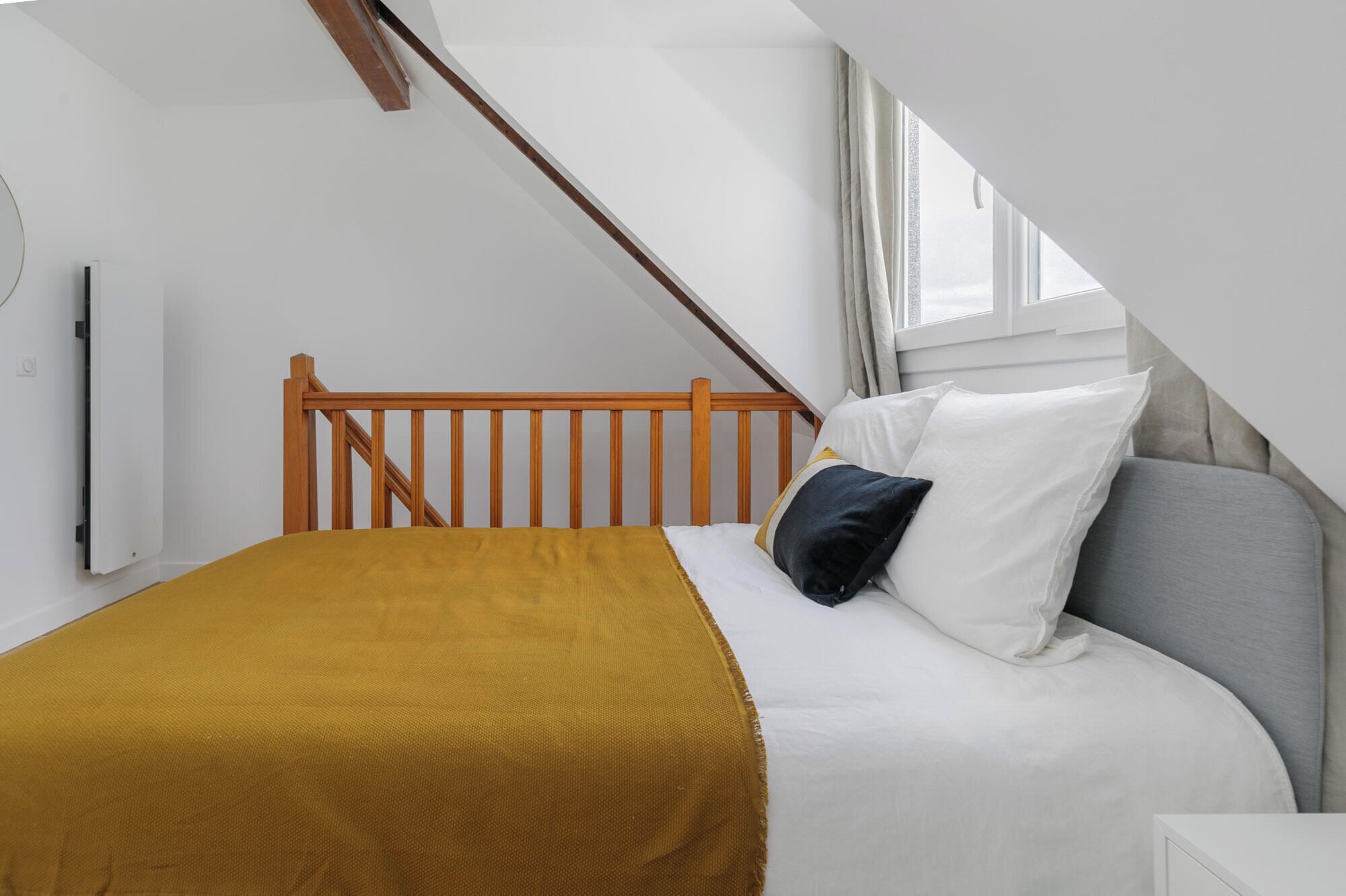 Une chambre à coucher avec une tête de lit en bois, un lit recouvert d’une literie blanche et jaune moutarde, et une fenêtre avec des rideaux.