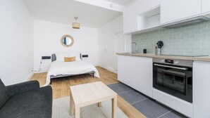 Ein kompakter, moderner Raum mit Bett, kleinem Tisch, Sofa, Küchenzeile inklusive Backofen und Spiegel an der Wand.