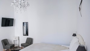 Un dormitorio con una cama grande, dos sillones, una mesita de noche pequeña, un televisor montado en la pared y una araña.