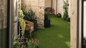Vista a través de una ventana de un patio con un pequeño césped, una silla de mimbre y plantas en macetas.