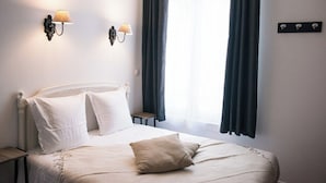 Ein ordentlich bezogenes Bett mit weißen Leinen und einem beigen Kopfkissen, zwei Wandlampen und ein Fenster mit dunklen Vorhängen.