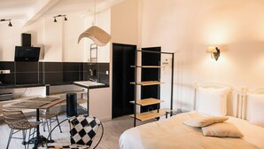 A modern hotel room with a bed, a dining area with chairs, a kitchen area with a TV, and a shelf unit. - Suite #4 - a unique 30m2 loft overlooking the inner courtyard (Aigues-Mortes)