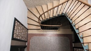 Eine Wendeltreppe mit Holzgeländer und beigefarbenen Stufen, die zu einem dunklen Zwischenpodest mit einer strukturierten Wand führt.