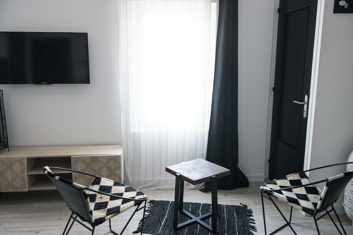 Een moderne kamer met een flatscreen televisie aan de muur, een klein houten tafeltje, twee geruitte fauteuils en een zwarte deur.
