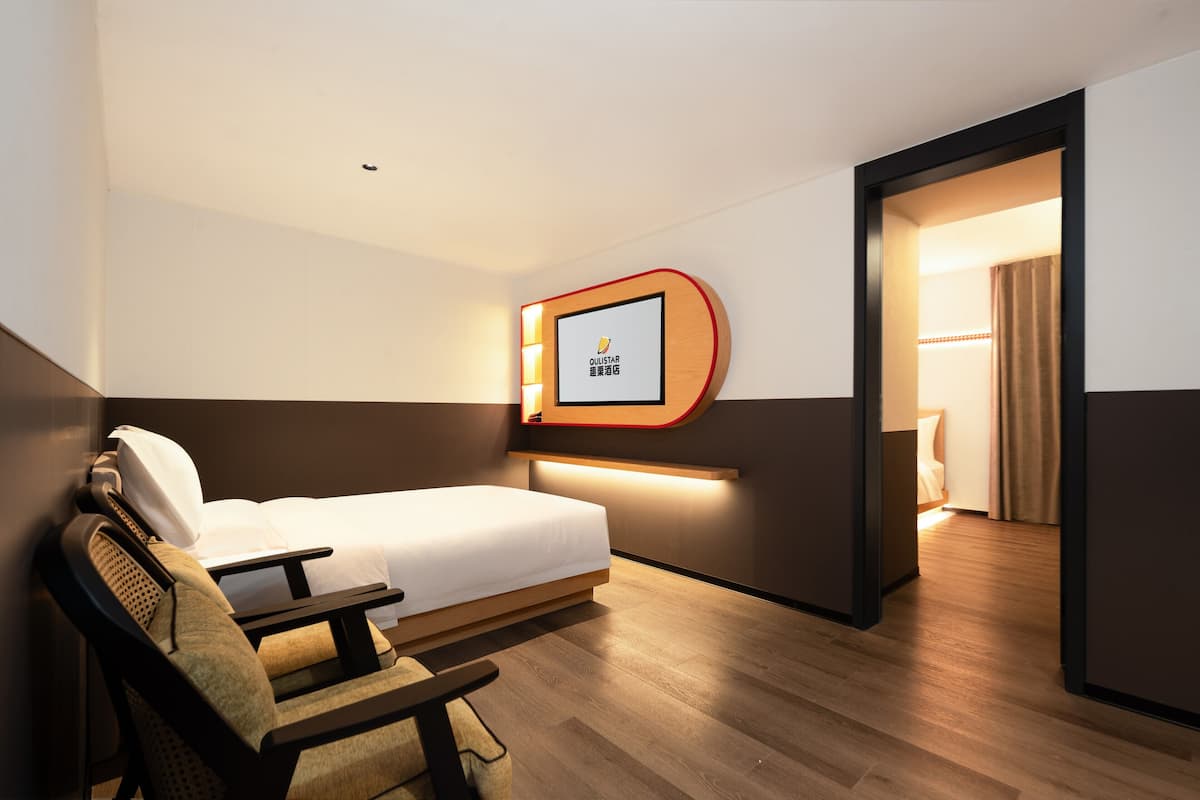 A hotel room with a bed, a desk, a chair, a wall-mounted TV, and a door leading to another room.