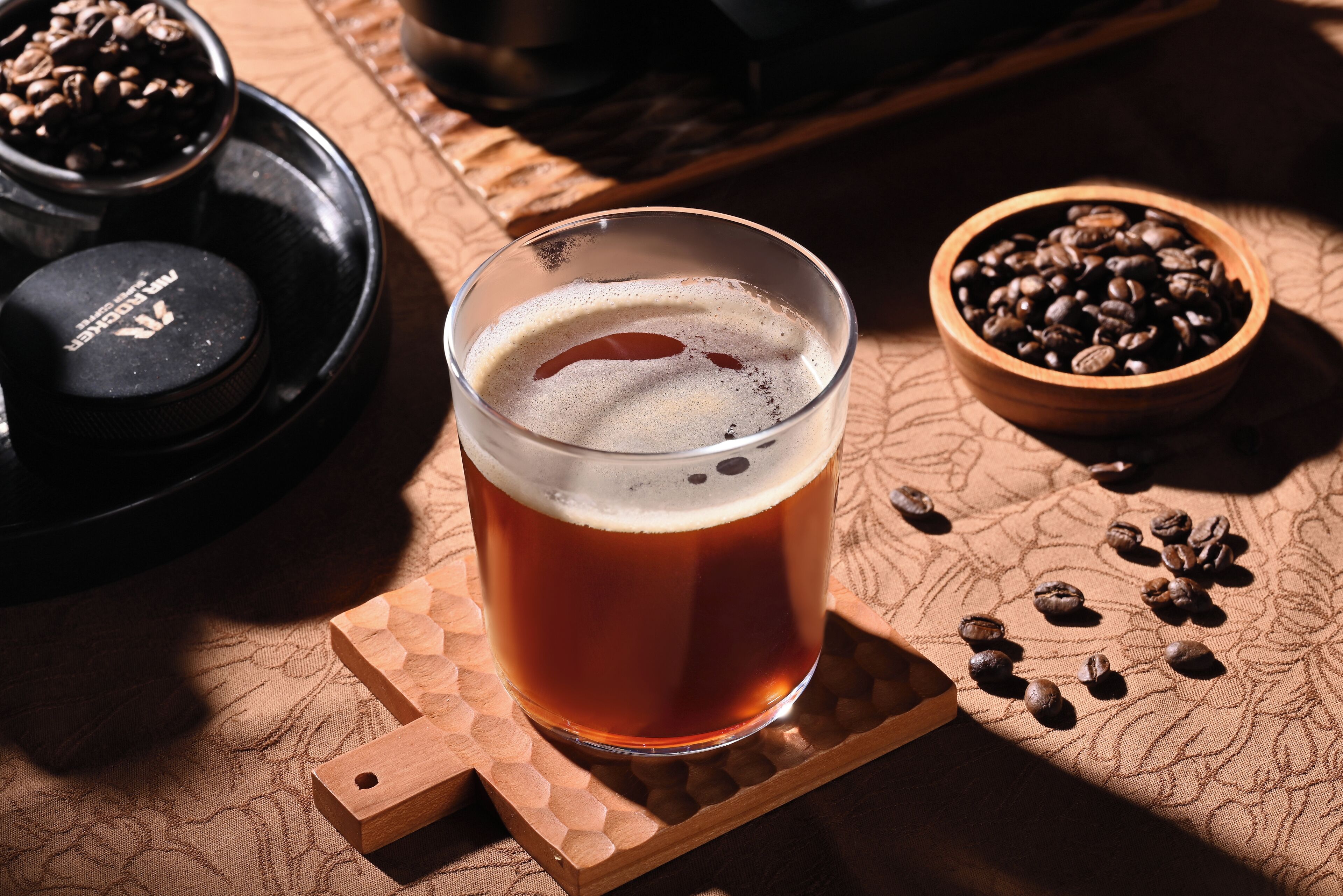A glass of amber beer on a wooden coaster, surrounded by coffee beans and a coffee grinder.