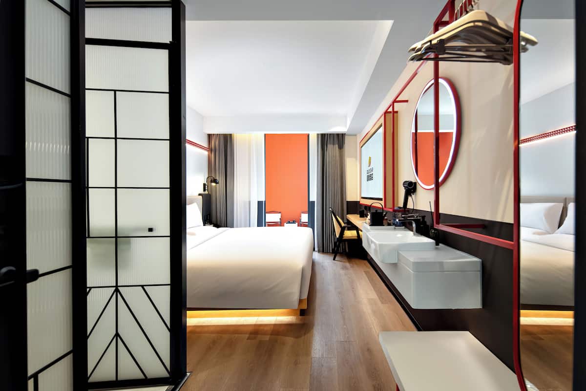 A modern hotel room with a bed, a desk, a chair, a large mirror, and a vanity with a sink.