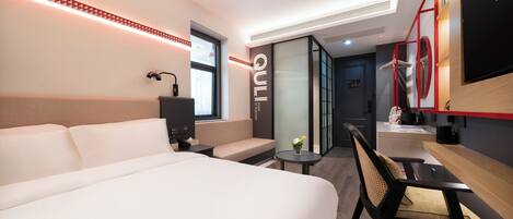 A modern hotel room with a large bed, a seating area, a desk, and a TV mounted on the wall.