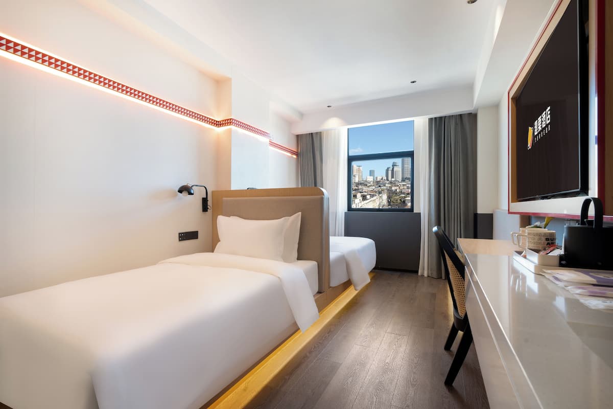 A hotel room with two beds, a desk, and a large window offering a city view.
