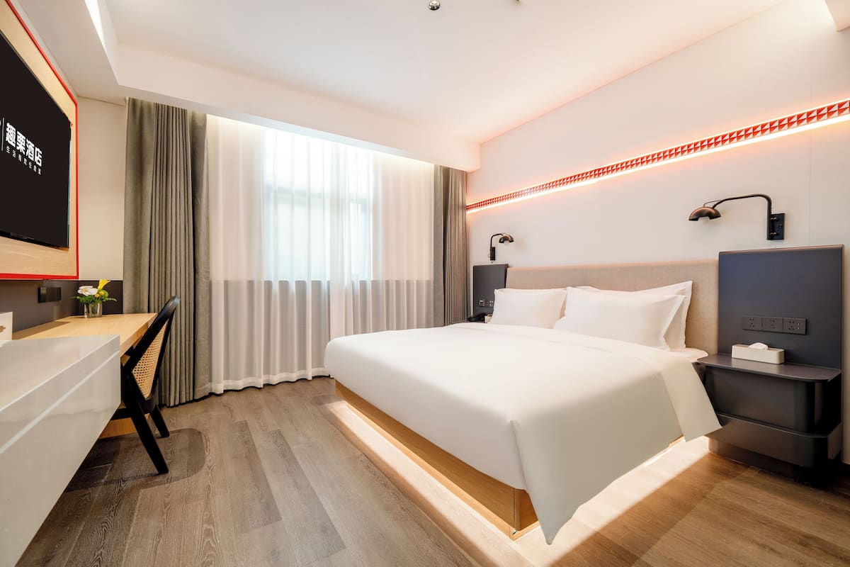 A modern hotel room with a large bed, a desk, and a chair. There is a window with curtains, a wall-mounted lamp, and a red LED light strip.