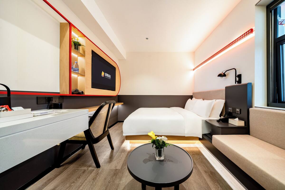 A modern hotel room with a bed, a desk, a chair, a small table with a vase, and a TV.