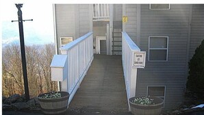 En sti der fører til en bygning med et fareskilt om glatte forhold. Der er to store plantekasser med blomster ved indgangen.