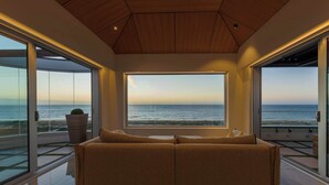 A modern living room with a large window overlooking the ocean, a wooden ceiling, and a beige sofa.