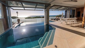 A modern infinity pool with clear blue water, a glass-enclosed deck, and a dining area with outdoor chairs.