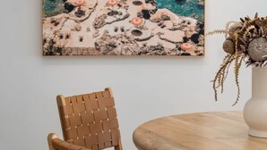 Een eetruimte met een ronde houten tafel, een geweven stoel en een schilderij van een kustlandschap aan de muur.