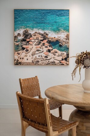 Un coin repas avec une table ronde en bois, une chaise en osier et un tableau représentant un paysage côtier accroché au mur.