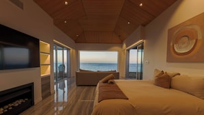 A modern bedroom with a large bed, a TV, a fireplace, and a view of the sea.
